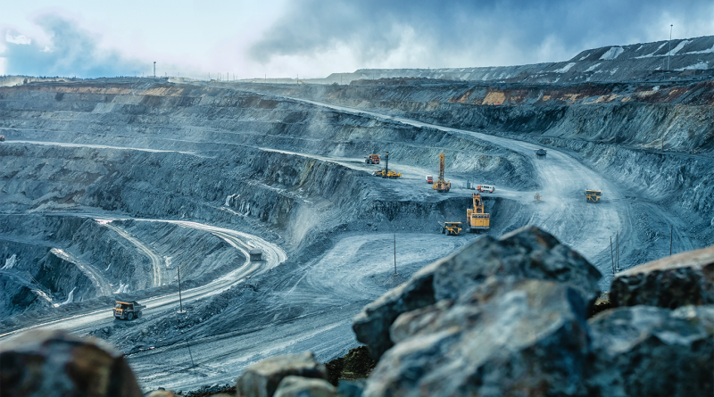 Large open-pit mining site with terraced excavation levels, haul trucks and drilling equipment operating in a dusty, overcast environment
