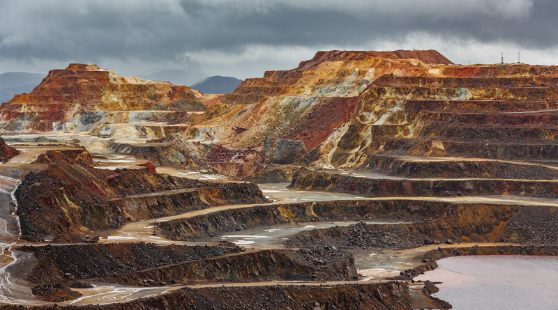Large open-pit mine with terraced excavation benches, winding haul roads and exposed mineral-rich rock formations beneath dark storm clouds, illustrating large-scale copper mining operations.